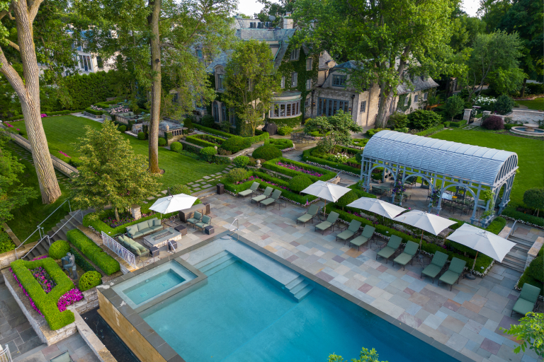 Aerial view of a luxurious backyard with a large swimming pool, manicured gardens, a pergola, and an elegant stone house surrounded by trees.