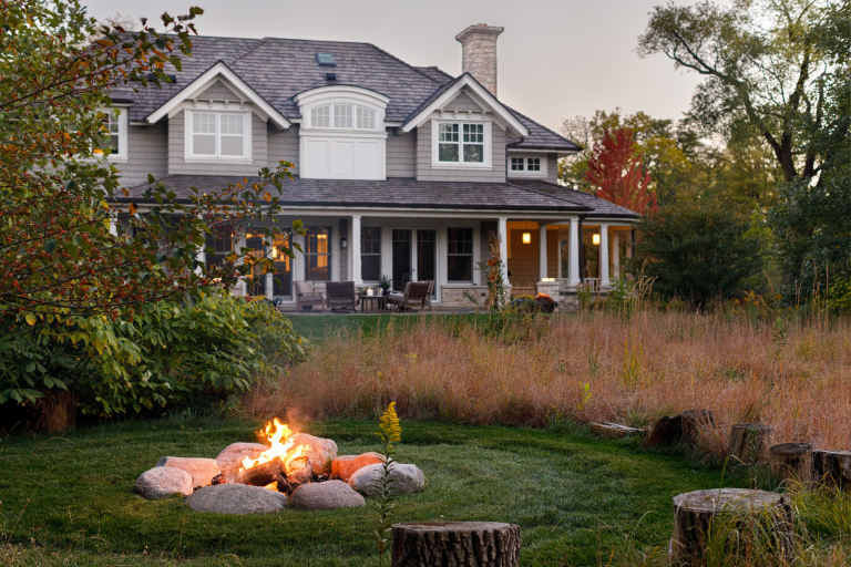 A cozy suburban house with an attached porch, surrounded by lush greenery and a lit fire pit in the foreground during twilight.