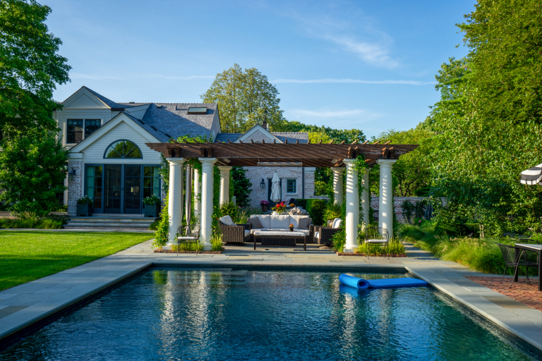 Luxurious backyard with a swimming pool, pergola with sitting area, and a large house surrounded by lush greenery.