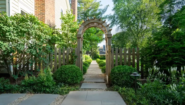 Wooden archway at the entrance of a garden path surrounded by lush greenery and shrubs, leading to a neatly landscaped yard.