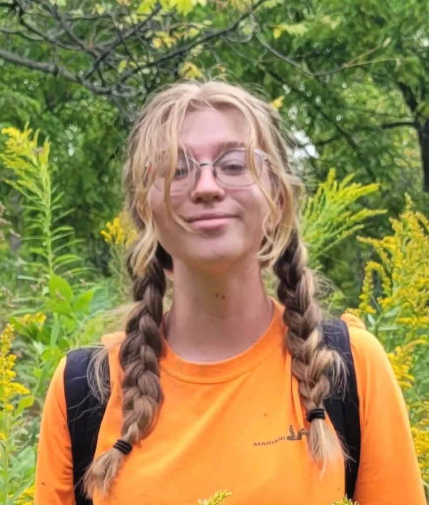 Person with glasses and braided hair, wearing an orange shirt and backpack, standing outdoors surrounded by green foliage and yellow wildflowers.