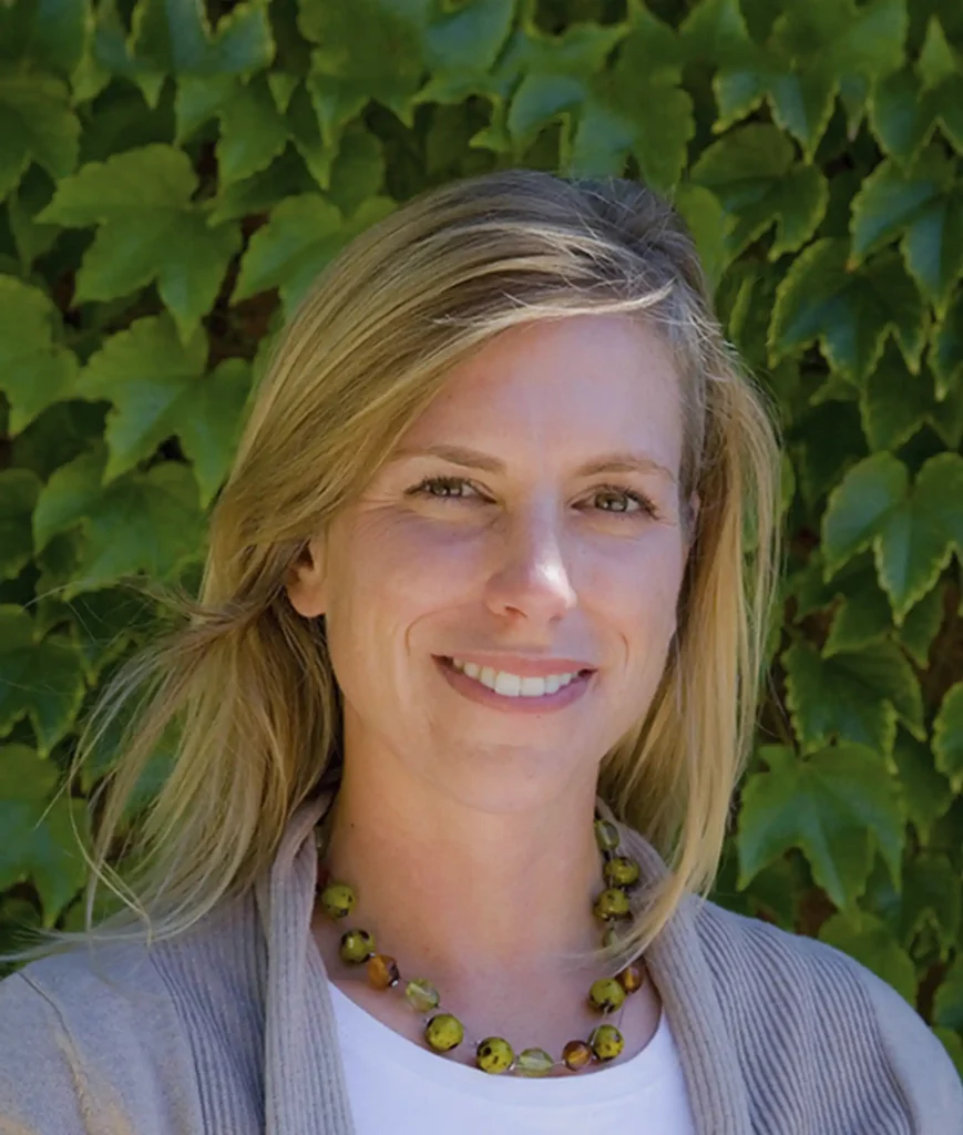 A woman with blonde hair smiles in front of a background of green ivy leaves. She is wearing a beige cardigan, white top, and a beaded necklace.