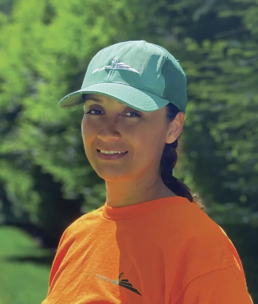 Person wearing an orange shirt and green cap, standing outdoors with greenery in the background.