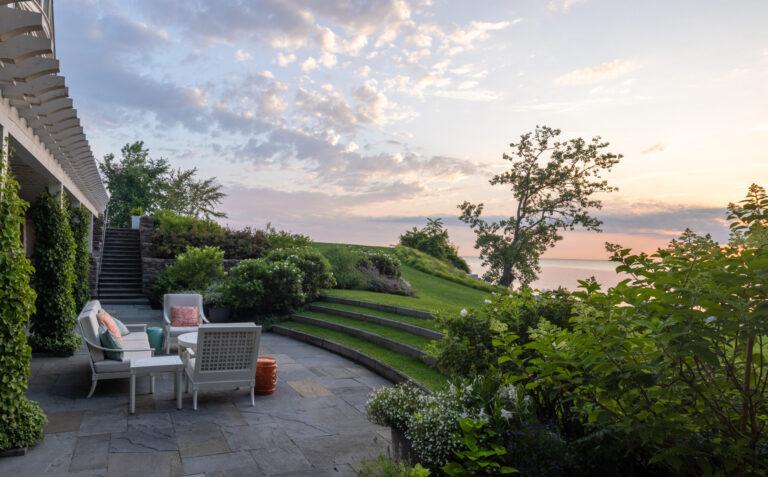 A scenic outdoor patio with cushioned furniture, surrounded by lush greenery, overlooks a set of stone steps leading to a grassy hill and a serene sunset view over a body of water—a perfect example of stunning landscaping in New Buffalo.