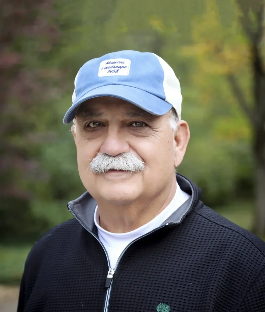 A man with a mustache wearing a blue and white cap, a white T-shirt, and a black zip-up jacket, standing outdoors.