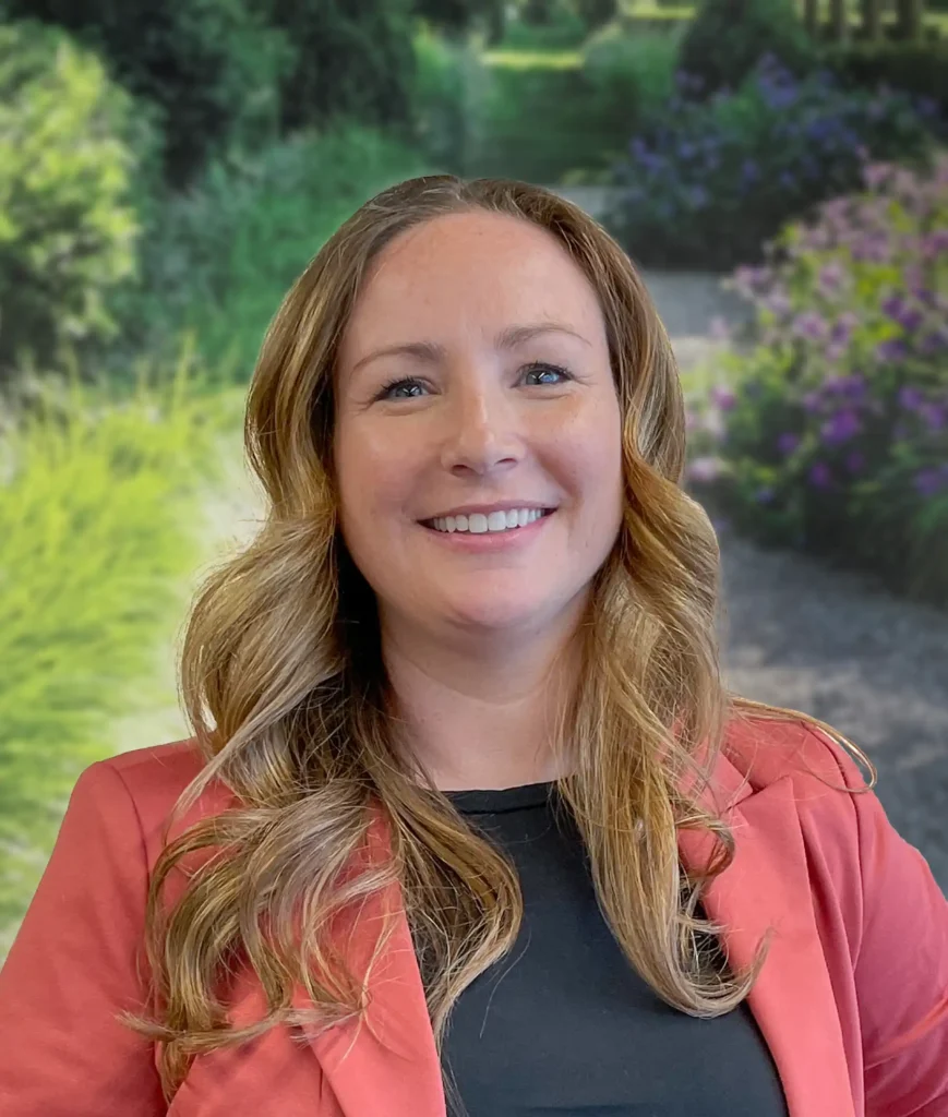 A woman with long wavy hair wearing a red blazer smiles in an outdoor garden setting with greenery and flowers in the background.