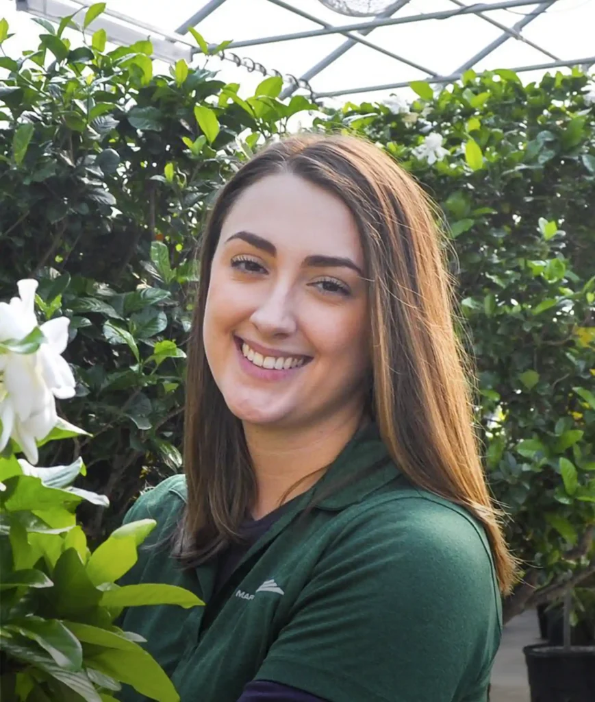 A woman with straight brown hair, wearing a green shirt, smiles while standing near lush green plants and a blooming white flower in a greenhouse.