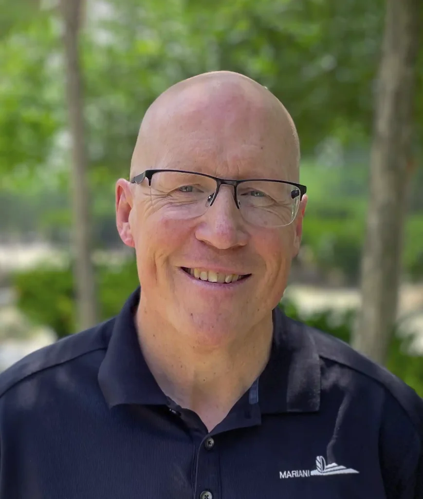 A man with glasses and a bald head is smiling while standing outdoors. He is wearing a dark polo shirt with a logo that says "Mariani". Trees and greenery are visible in the background.