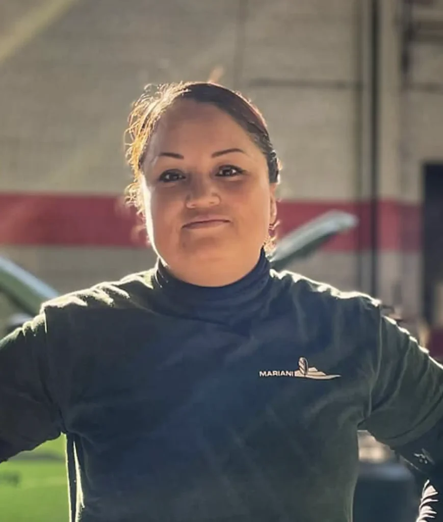 A person in a dark green shirt stands confidently with hands on hips, in a well-lit automotive workshop with a car in the background.