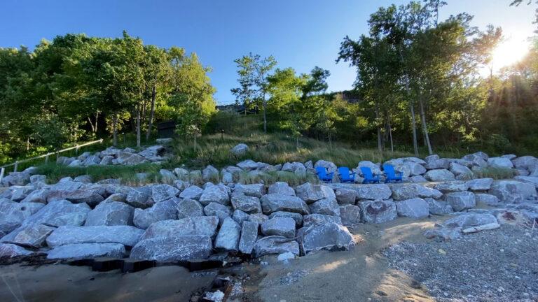 A rocky shoreline with several blue chairs arranged among large rocks showcases the beautiful landscaping in New Buffalo. Trees and greenery extend up a hill in the background under a clear sky, creating a serene and picturesque view.