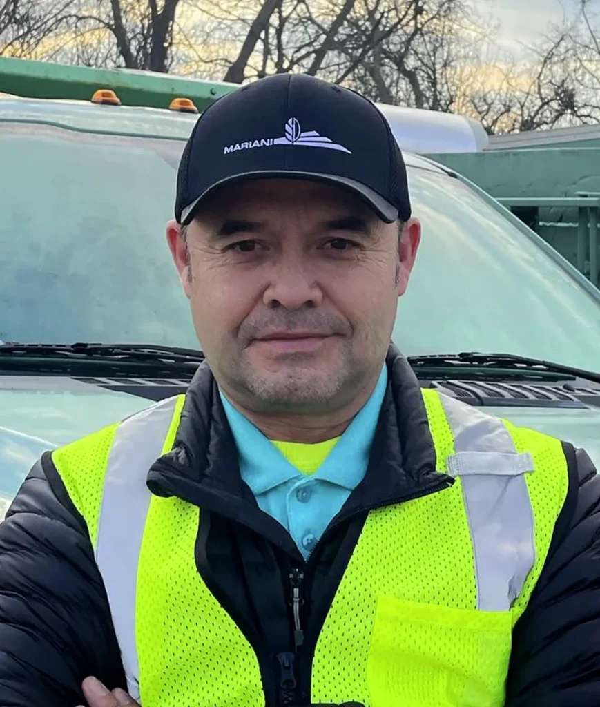 Man in high-visibility jacket and baseball cap stands outdoors in front of a truck, with trees and sky in the background.