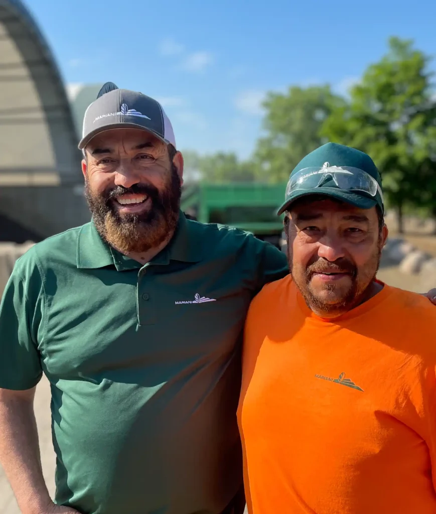 Two men are posing together outdoors. One wears a green polo shirt and cap, while the other wears an orange shirt and green cap. Both shirts have a small logo. A clear, bright sky and trees are behind them.