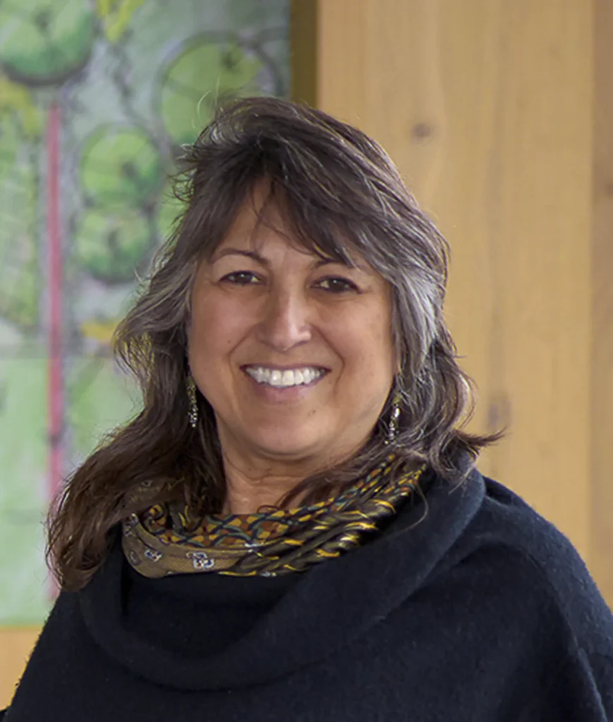 A smiling person with long, graying hair wearing a dark top and patterned scarf stands indoors with a blurred background.