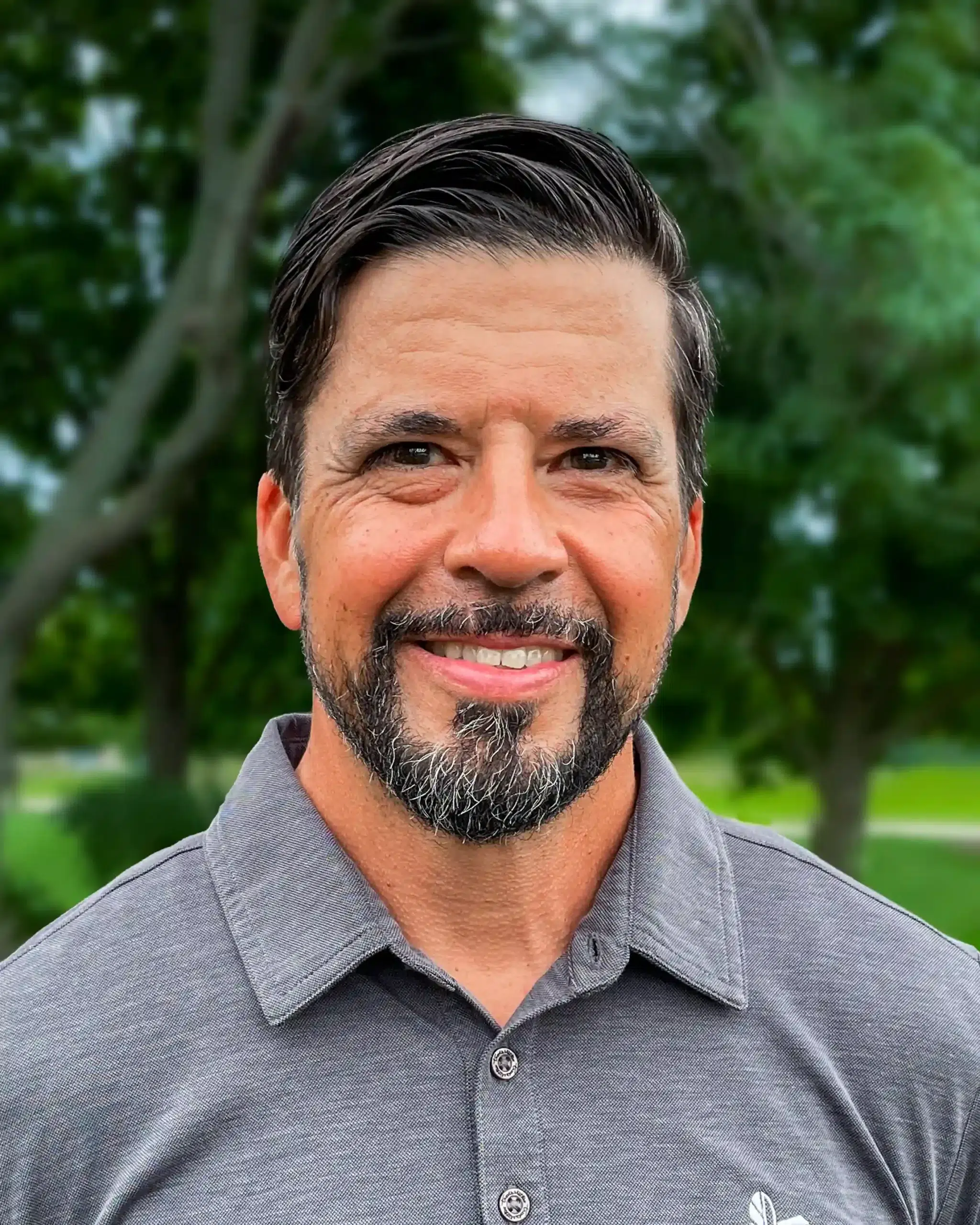 A man with short dark hair and a beard, wearing a grey polo shirt, smiles in an outdoor setting with trees in the background.