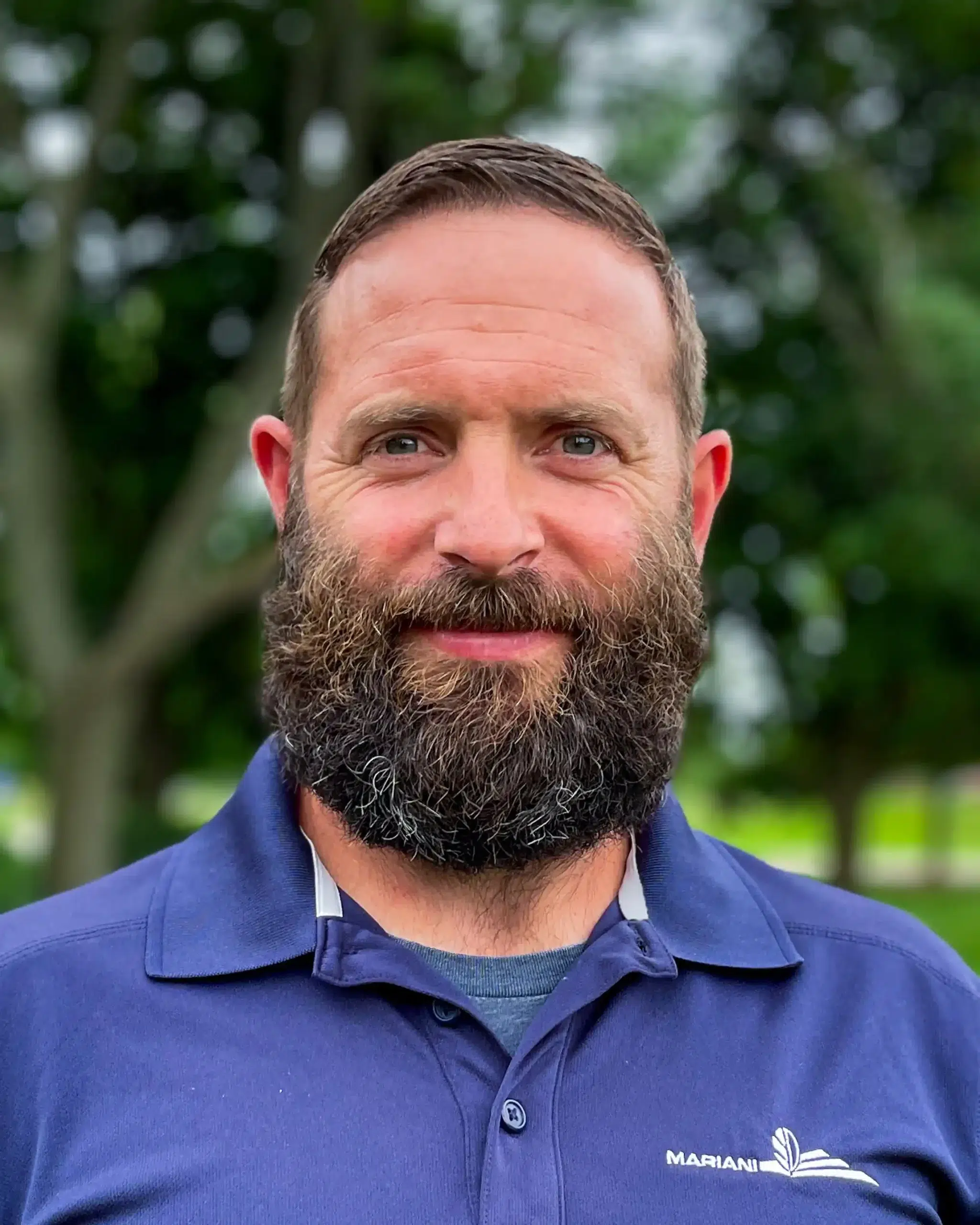 A man with a beard and short hair wearing a navy polo shirt stands outdoors, looking at the camera. Trees are visible in the blurred background.