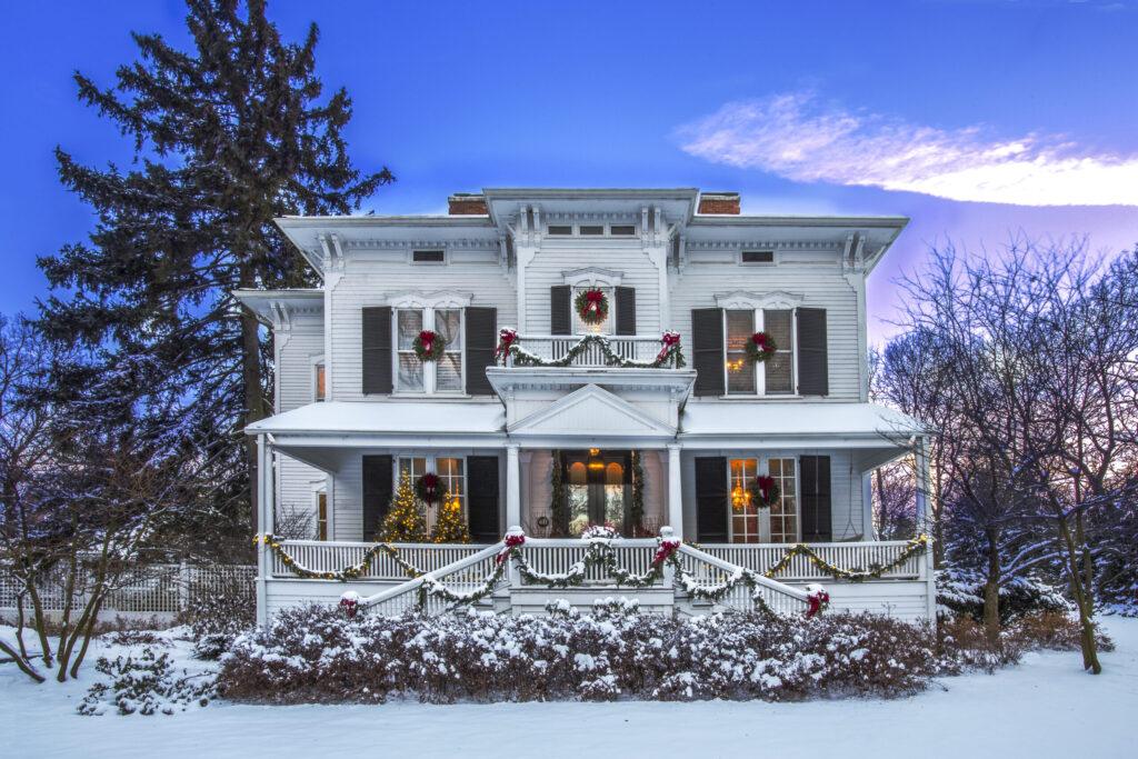 A two-story house decorated with Christmas wreaths and garlands, surrounded by a light layer of snow, with a tall tree on the left and bare trees on the right at sunset.