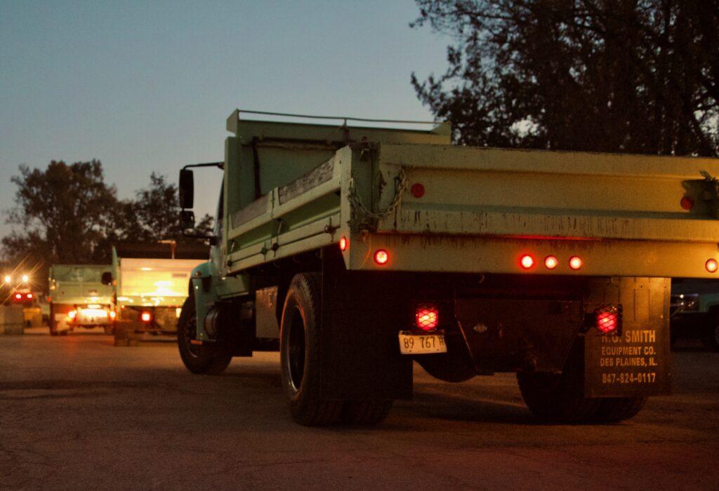 Two green dump trucks, integral to commercial landscape construction, are parked on a road at dusk, their brake lights glowing red against the fading light. Tall trees frame this serene scene in the background.