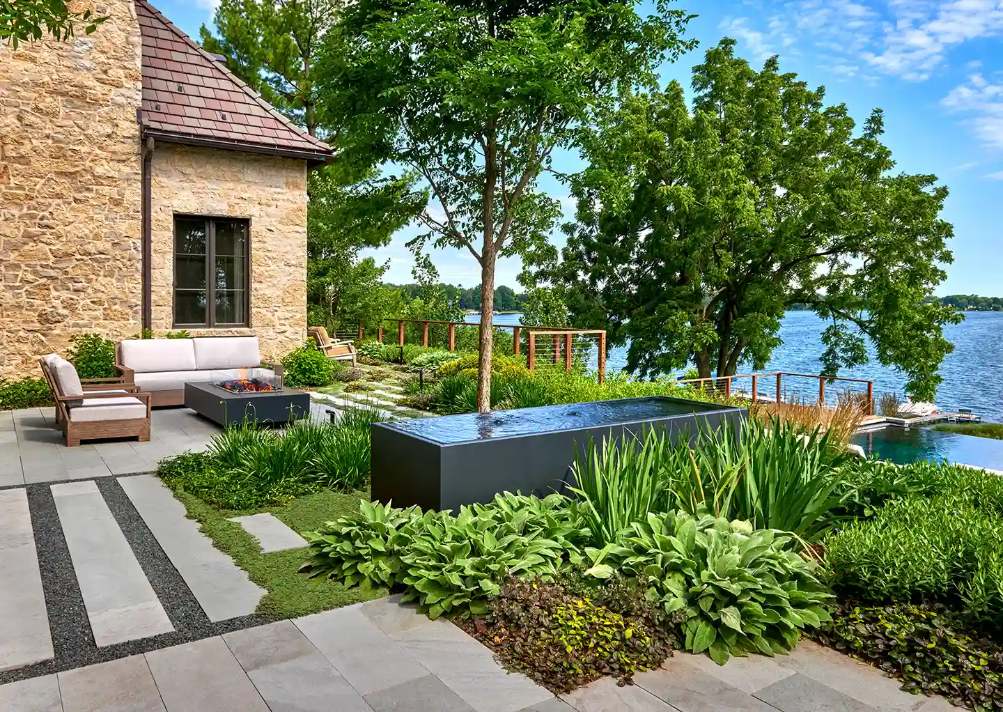 A landscaped backyard with patio furniture, lush greenery, a water feature, and a view of a lake surrounded by trees under a blue sky.