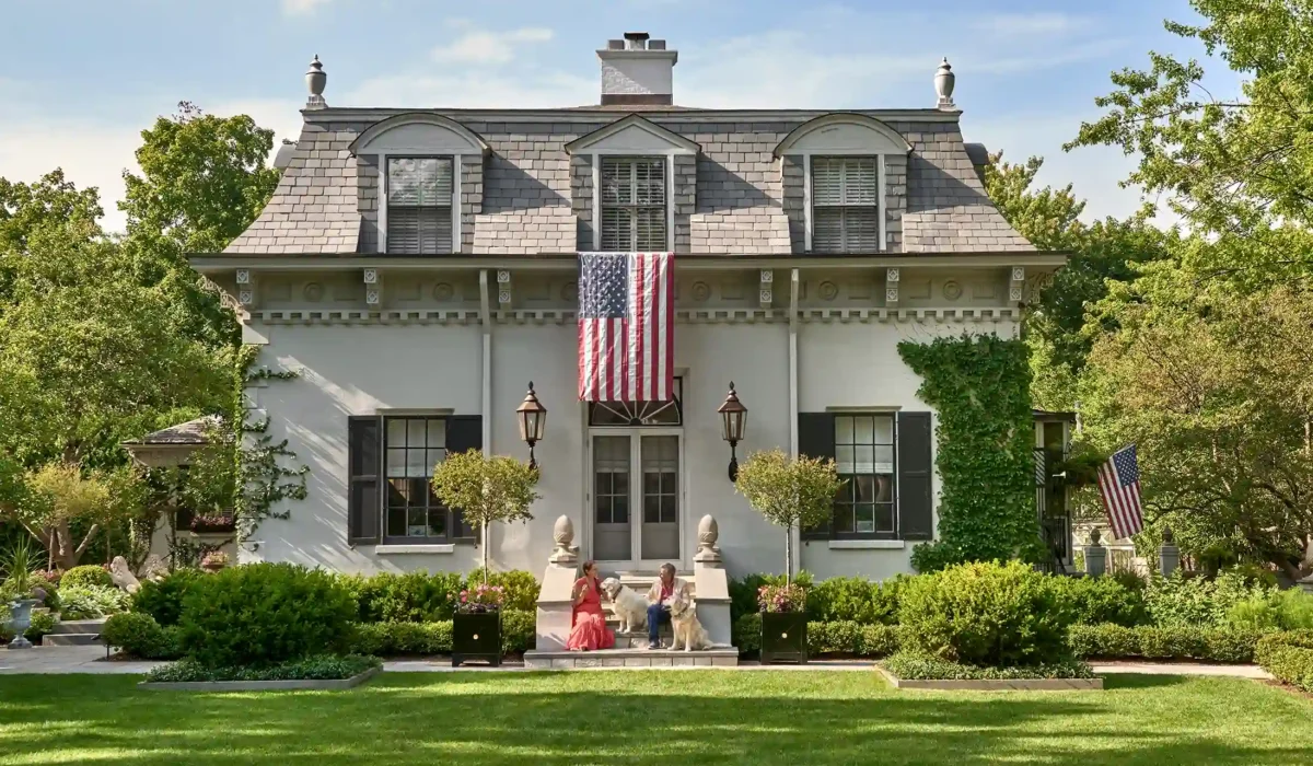 A stately two-story house with a gray roof, dormer windows, and an American flag. A manicured lawn and lush greenery surround the home, with two people sitting on the front steps.