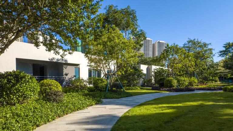 The pathway meanders through a meticulously maintained garden featuring lush grass, vibrant shrubs, and towering trees, all under the watchful care of Landscape Maintenance for HOAs. This tranquil scene is bordered by modern white buildings with a backdrop of tall structures against a clear blue sky.